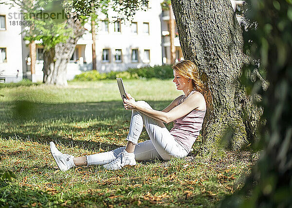 Lächelnde Frau mit Tablet-PC sitzt an einem sonnigen Tag am Baumstamm im Park