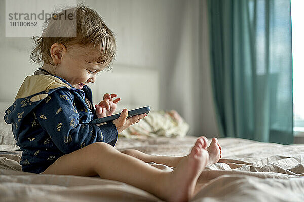 Glücklicher kleiner Junge  der im Schlafzimmer ein Spiel auf dem Smartphone spielt