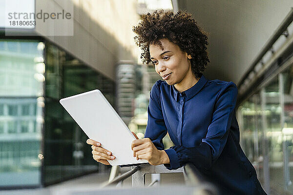 Lächelnde Geschäftsfrau beobachtet Tablet-PC  der am Geländer lehnt