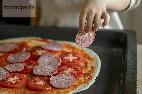 Hand of boy preparing pizza with pepperoni in kitchen at home