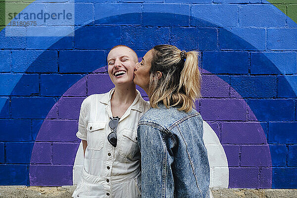 Happy woman kissing on cheeks of non-binary friend in front of colorful wall