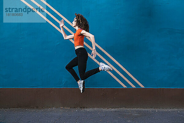 Junge Frau springt auf Fußweg vor blauer Wand