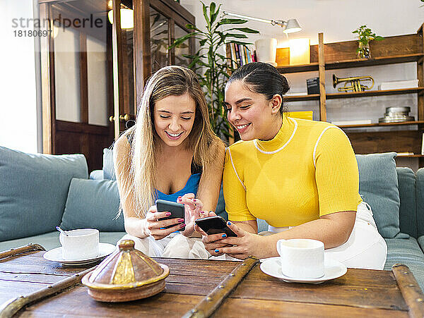 Lächelnde junge Frauen  die zu Hause auf dem Sofa Mobiltelefone teilen