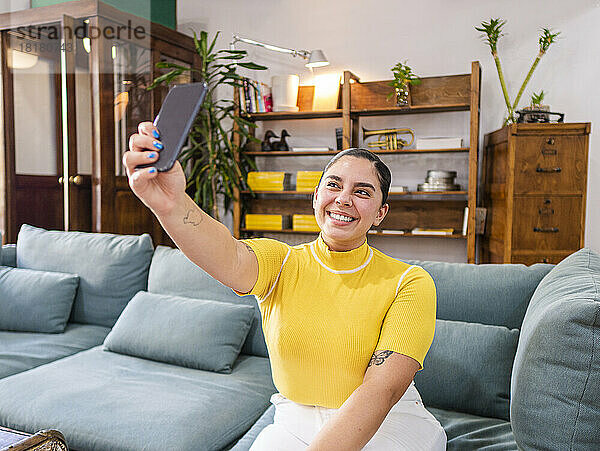 Lächelnde junge Frau  die zu Hause auf dem Sofa ein Selfie mit dem Smartphone macht