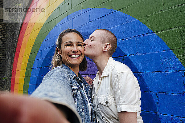 Nicht-binäre Person küsst Freund vor Regenbogenwand
