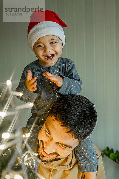 Netter Junge sitzt auf der Schulter seines Vaters und schmückt zu Hause den Weihnachtsbaum
