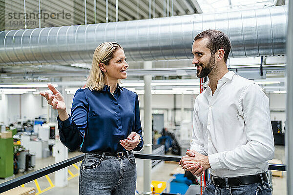 Lächelnde Geschäftsfrau gestikuliert und spricht mit einem Kollegen am Geländer der Fabrik