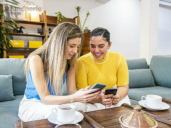 Happy young women sharing mobile phones on sofa at home