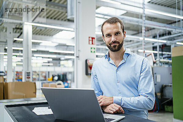 Lächelnder Geschäftsmann mit Laptop auf dem Tisch in der Fabrik