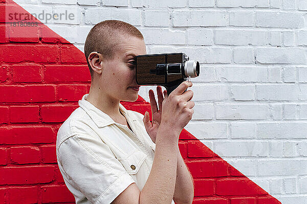 Nicht-binäre Person hält Videokamera vor zweifarbiger Wand