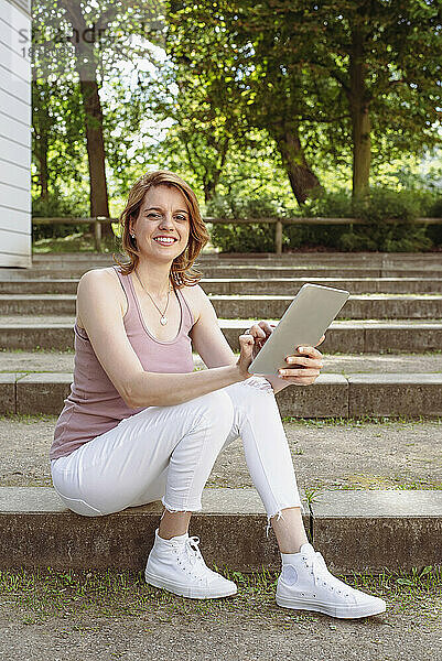 Glückliche Frau mit Tablet-PC sitzt auf der Treppe im Park