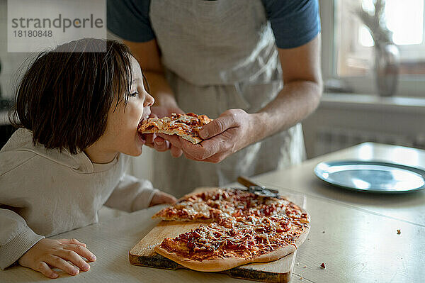Man feeding pizza to son in kitchen at home
