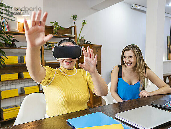 Lächelnde Geschäftsfrau gestikuliert mit VR-Brille  die neben einem Freund am Tisch sitzt