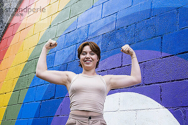 Glückliche junge Frau  die vor einer an die Wand gemalten Regenbogenfahne ihre Muskeln spielen lässt