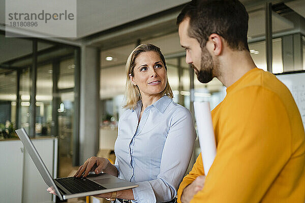 Lächelnde Geschäftsfrau mit Laptop und Blick auf Kollegen im Büro