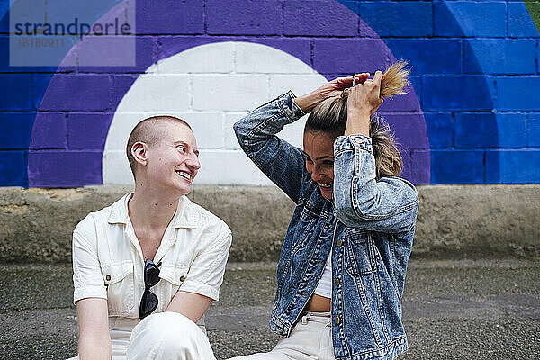 Glückliche Frau bindet Haare mit nicht-binärer Person  die auf einem Fußweg sitzt