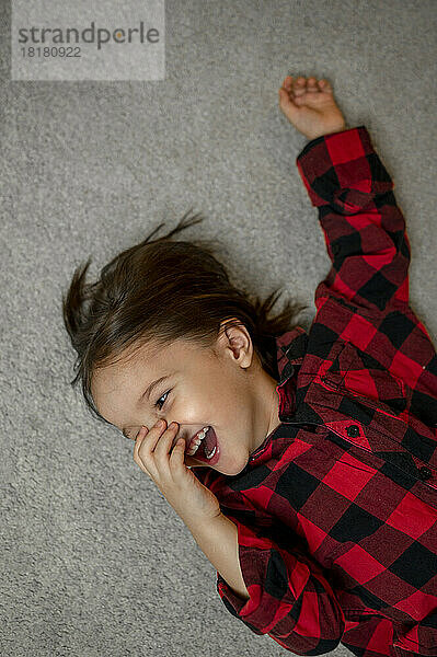 Fröhlicher Junge trägt kariertes Hemd und liegt auf dem Teppich