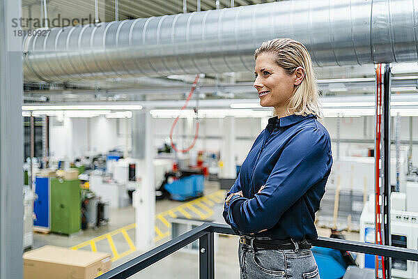 Lächelnde Geschäftsfrau steht mit verschränkten Armen neben dem Geländer in der Fabrik