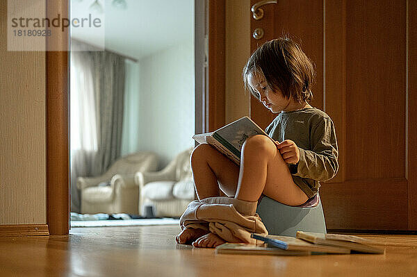 Netter Junge sitzt auf dem Töpfchen und liest zu Hause ein Buch