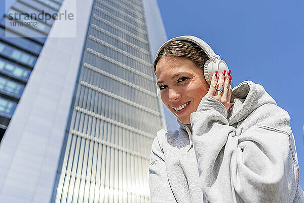 Lächelnde Frau  die an einem sonnigen Tag vor dem Gebäude Musik über drahtlose Kopfhörer hört