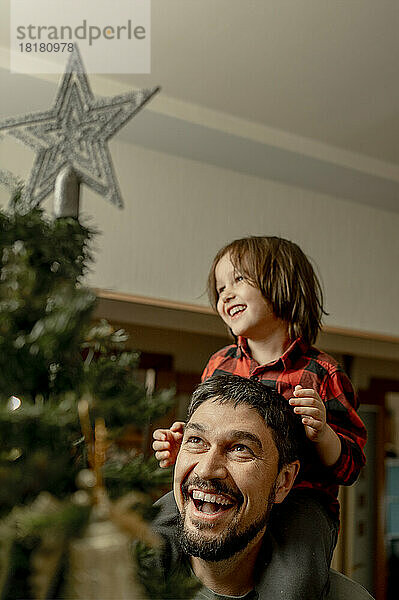 Glücklicher Mann trägt Sohn auf Schultern und schaut auf den Weihnachtsbaum