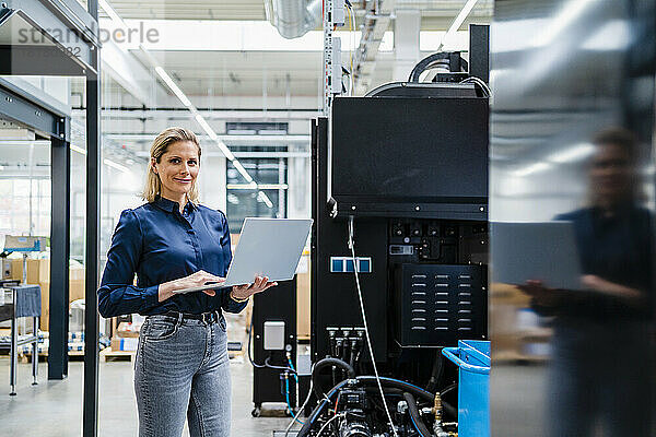 Lächelnde Geschäftsfrau mit Laptop steht neben Maschinen in der Industrie