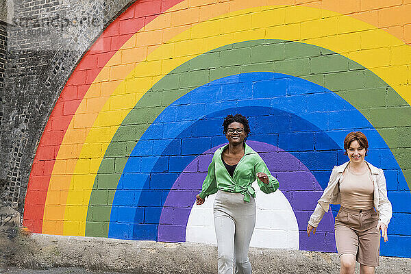 Glückliche Frau mit Freundin läuft vor regenbogenfarbener Wand