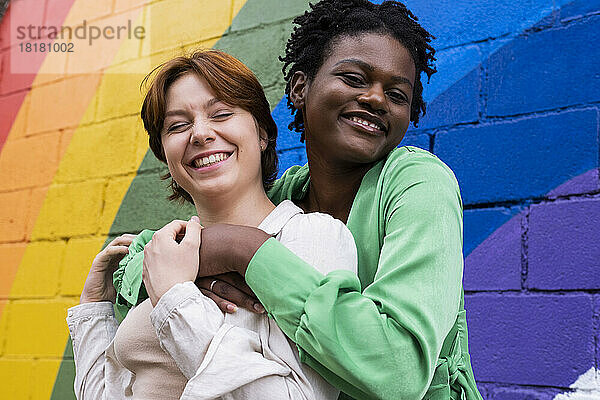 Glückliche Frau umarmt Freundin vor einer an die Wand gemalten Regenbogenfahne