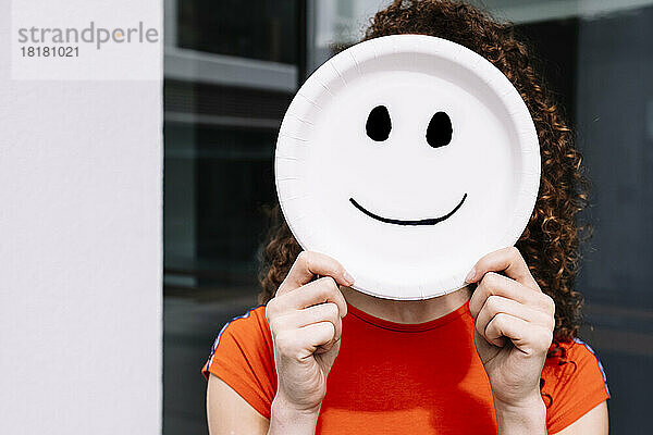 Junge Frau hält lächelndes Emoticon-Teller über Gesicht
