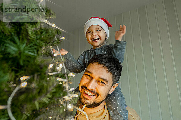 Lächelnder Junge sitzt auf Vaters Schulter und hat Spaß beim Schmücken des Weihnachtsbaums zu Hause