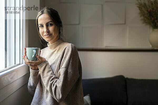 Lächelnde junge Frau mit einer Tasse Tee  die zu Hause am Fenster steht