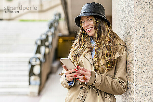 Lächelnde Geschäftsfrau mit Hut und Mobiltelefon an der Wand