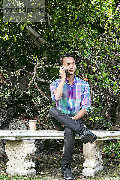 Mann sitzt mit Kaffeetasse im Park und telefoniert mit Smartphone
