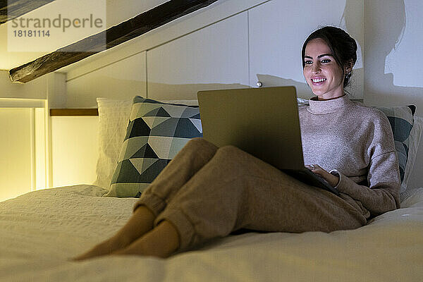 Glückliche junge Frau benutzt Laptop im Schlafzimmer