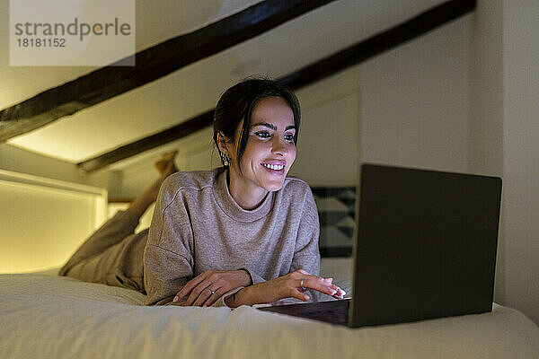 Lächelnde junge Frau benutzt Laptop auf dem Bett im Schlafzimmer