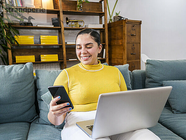 Lächelnde Geschäftsfrau benutzt Smartphone und sitzt mit Laptop auf dem Sofa