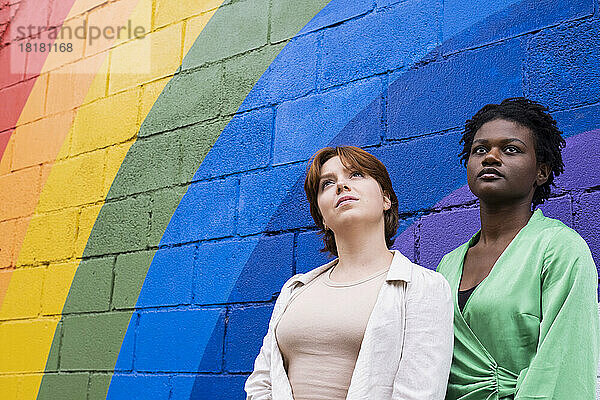 Nachdenkliche junge Frau mit Freundin vor regenbogenfarbener Wand