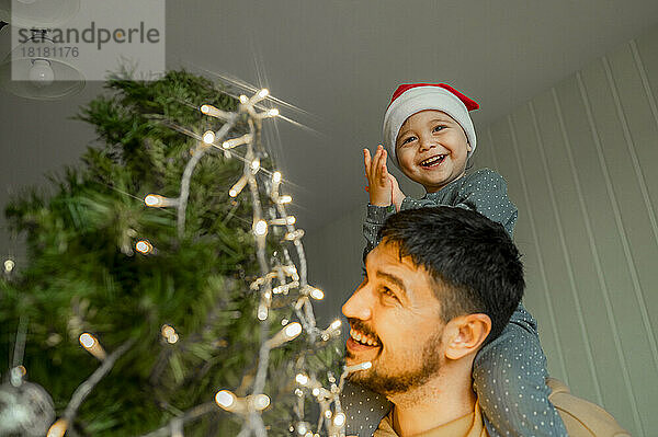 Lächelnder Junge sitzt zu Hause auf Vaters Schulter am Weihnachtsbaum