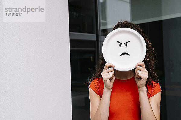Junge Frau hält wütendes Emoticon-Teller über Gesicht