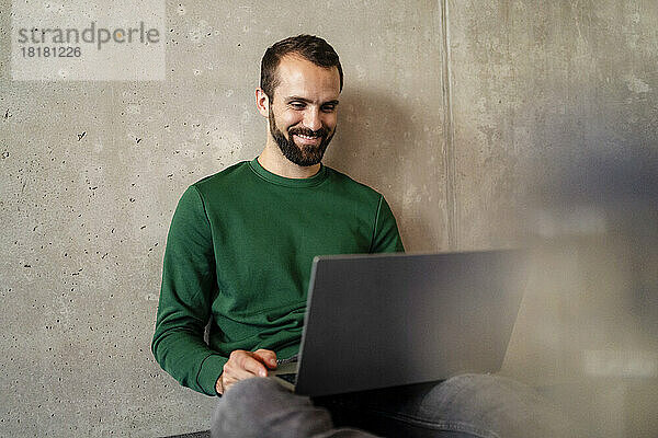 Glücklicher Geschäftsmann sitzt mit Laptop vor der Wand im Büro