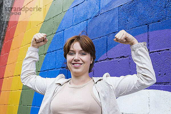 Lächelnde junge Frau  die vor einer an die Wand gemalten Regenbogenfahne ihre Muskeln spielen lässt