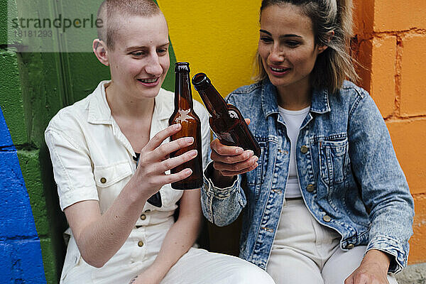Schwules Paar toastet Bierflaschen und sitzt vor einer bunten Wand