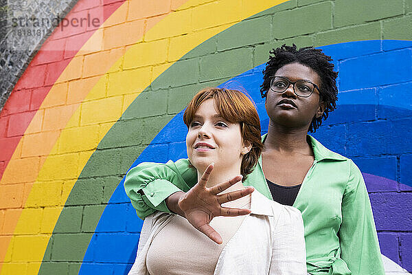 Frau umarmt Freundin von hinten vor regenbogenfarbener Wand