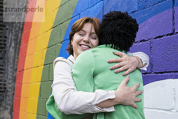 Lächelnde Frau umarmt Freundin vor einer an die Wand gemalten Regenbogenfahne
