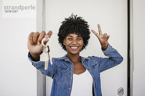 Glückliche Frau zeigt Hausschlüssel und Peace-Zeichen vor der Tür