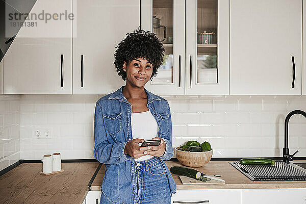 Lächelnde junge Frau mit Smartphone lehnt zu Hause an der Küchentheke