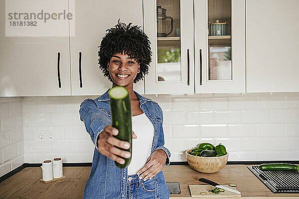 Glückliche junge Frau zeigt Zucchini und lehnt zu Hause an der Küchentheke