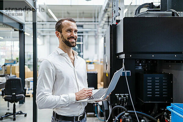 Lächelnder junger Geschäftsmann mit Laptop in der Nähe von Maschinen in der Fabrik