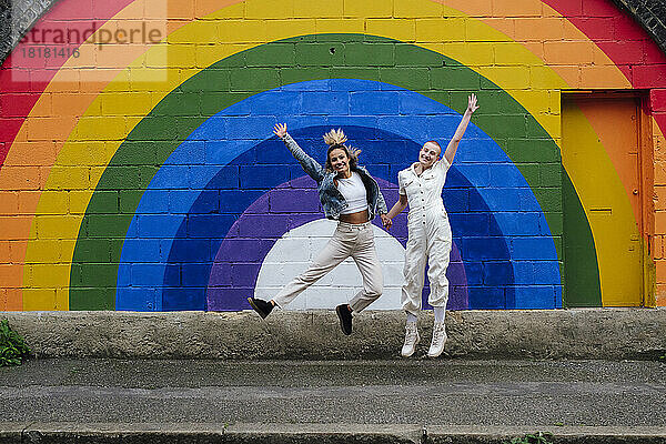 Glückliches lesbisches Paar springt vor einer regenbogenfarbenen Wand