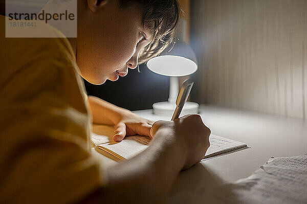 Junge schreibt zu Hause unter beleuchteter Lampe in ein Buch